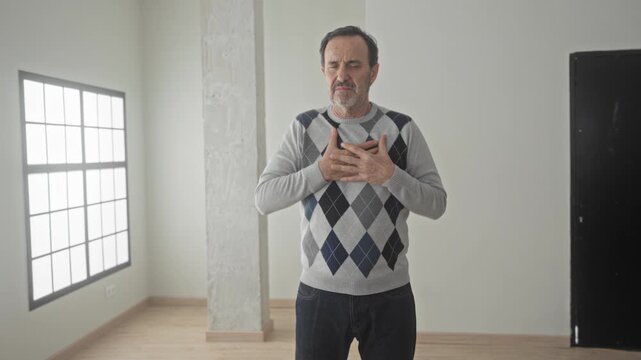 Man clutching hands to chest in building house interior by window wearing argyle sweater and jeans; heartattack pain.
