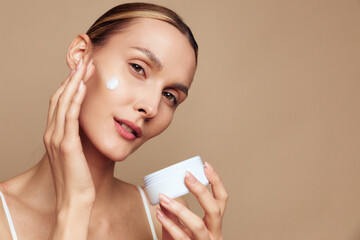 Woman applying face cream in a close up on a beige background, holding a white jar, soft studio lighting, skincare routine, wellness and self care, fresh healthy glow