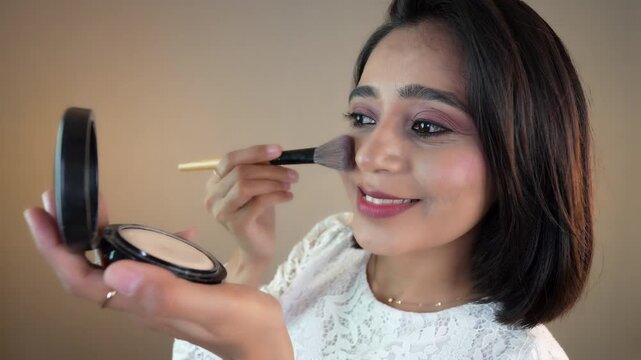 Close up of happy beautiful young Indian woman applying compact powder with brush to beautiful face, isolated over grey studio background.