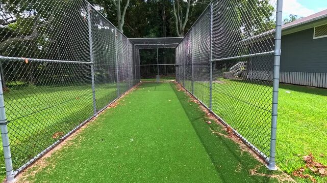 Walking through empty outdoor cricket net with green turf and chain-link fencing on sunny day.