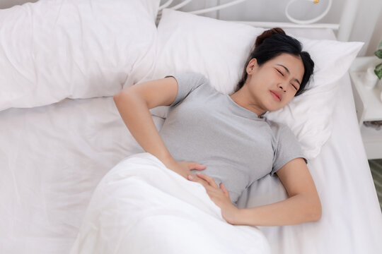 Asian women lying in bed holding stomach in pain, showing abdominal discomfort, menstrual cramps or digestive problem. Healthcare, illness, Gastritis and enteritis, copy space.