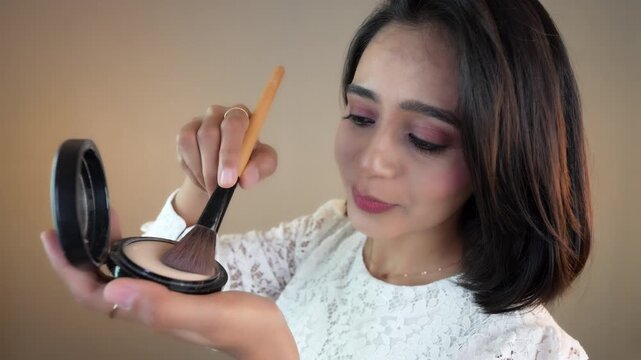 Close up of happy beautiful young Indian woman applying make up hold powder compact, isolated over grey studio background.