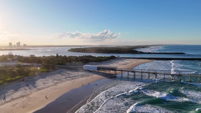 Aerial view of a long pier stretching into ocean with gentle waves and distant city skyline at sunset.