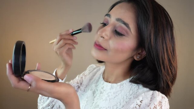 Close up of happy beautiful young Indian woman applying compact powder with brush to beautiful face, isolated over grey studio background.