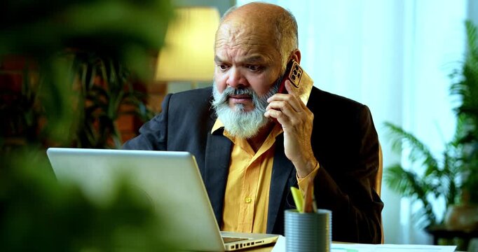Indian senior businessman or corporate executive multitasking by speaking on a smartphone while actively typing on a laptop at his desk in a cozy office with professional moody lighting.