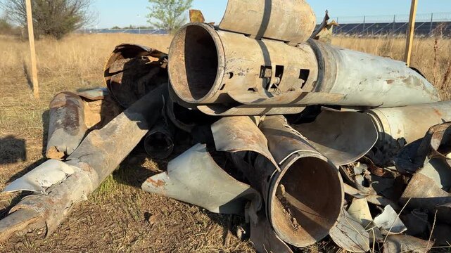Footage of destroyed metal pipes and rocket fragments scattered across a dry grassy field in a rural area. The scene shows heavily rusted cylindrical parts, torn metal edges