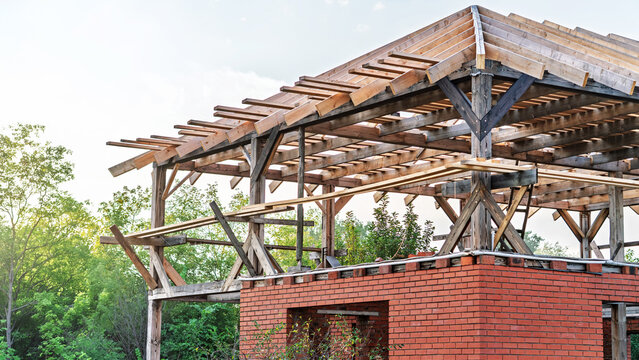 Red brick first floor, wooden frame exposed rafters, partial roof, open window voids, green foliage backdrop, soft cloudy sky, raw masonry work, site awaiting