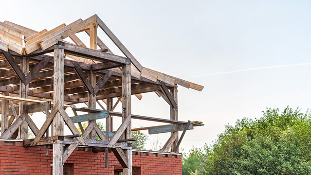 Unfinished house structure features a sturdy wooden frame and exposed roof trusses. Below, new red brick foundation walls are visible, surrounded by green trees under a bright daytime sky