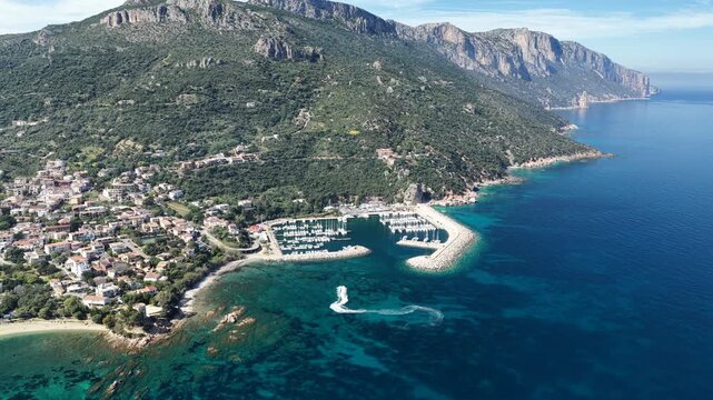 Aerial drone view of Santa Maria Navarrese, featuring turquoise waters, sandy beaches, marina harbor and dramatic limestone mountains along the eastern Sardinian coastline on a sunny day.