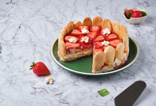 Round tiramisu cake with fresh strawberries and mascarpone cream decorated with marshmello flowers on a green plate on a light marble background. No-bake desserts.