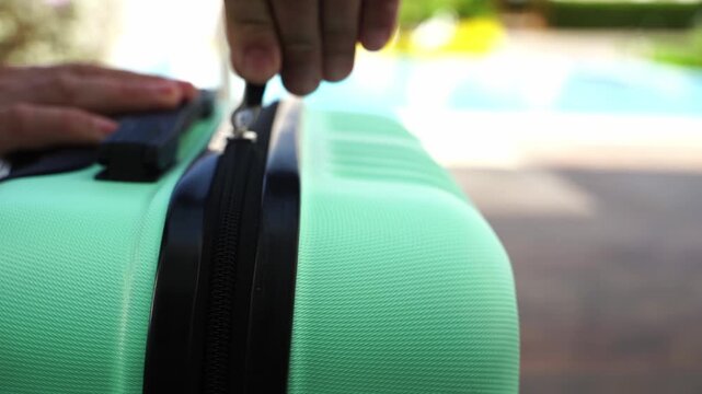Hand zips mint suitcase near pool. Gentle motion suggests travel or arrival. Bright colors imply vacation or leisure. Close-up captures everyday traveler's moment