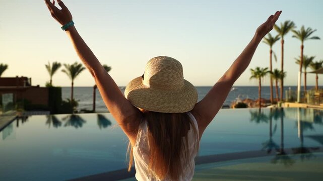 Sunset bathes a tranquil poolside scene. Woman in a wide-brimmed hat stands facing the sea. Her arms rise in joyful release. Palm trees frame the distant horizon. Gentle light reflects off water
