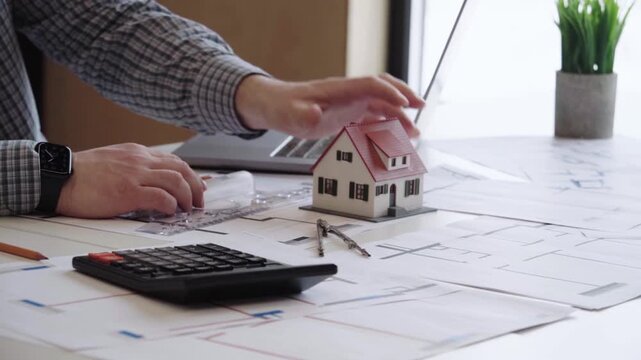 Man holds tiny house model beside blueprints. He checks calculations with a calculator. His watch glints as he works. Focused, calm, and thoughtful. Great for real estate, finance