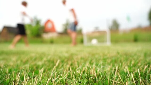 Grass blades fill the foreground sharply. Two figures move blurred in the distance. They play soccer in an open field. Sunlight illuminates the green expanse