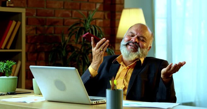 Indian senior businessman or corporate executive multitasking by speaking on speakerphone via his smartphone while actively typing on a laptop at his desk in a cozy office with moody lighting.