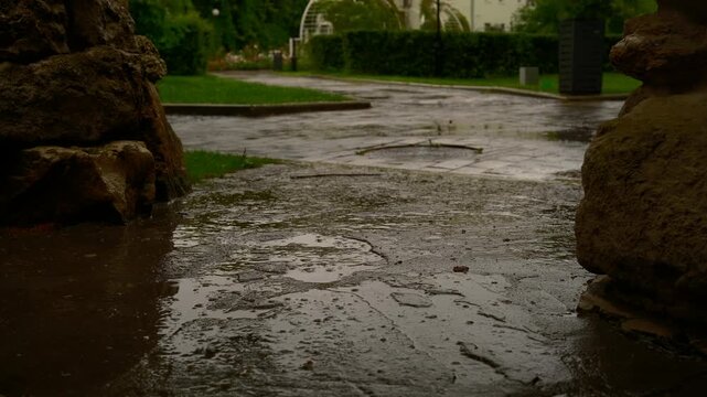 Cinematic video of summer rain falling on natural stone surfaces in a city park. The scene captures raindrops hitting textured stone walls and rock elements, creating a calm and atmospheric