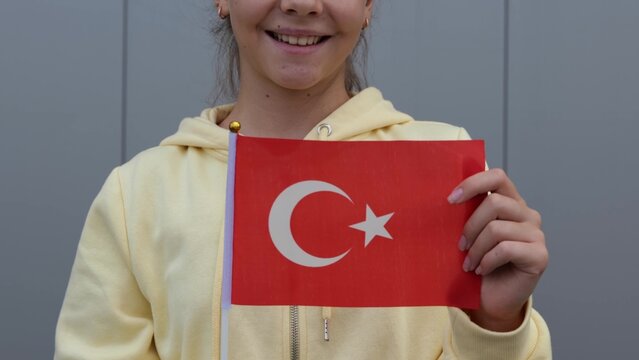 Caucasian teenage Girl waving small flag of Turkey. Zoom out, close up, grey background, female small girl in yellow hoodie, keeping flag of Turkey in her hands.