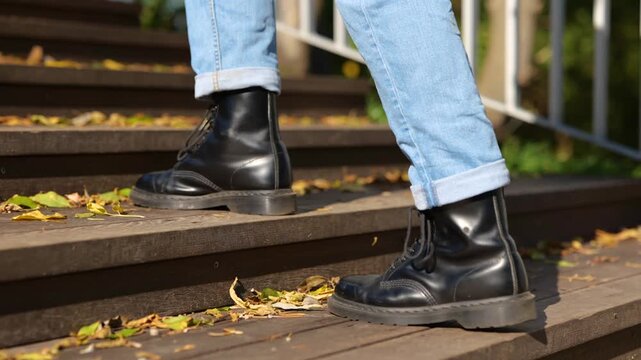 Guy in black boots going up the stairs in the park during sunny day. Leaves on the stairway. Cropped. Concept of autumn walks. Slow motion