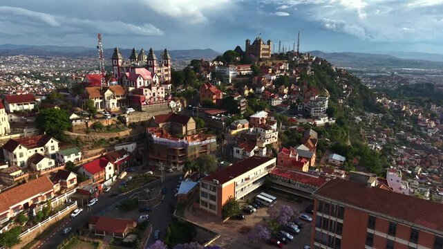 Aerial views capture vibrant hillside town with historic church and colorful homes. Purple flowering trees add vivid contrast to red rooftops. City sprawls below under cloudy skies