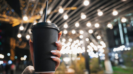 Close-up of a black paper coffee cup in women's hands against the background of a winter street with lights. © Anastasia