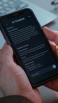 Close up of hands holding smartphone with AI chatbot application at workspace with laptop. Artificial intelligence assistant app helps improving business productivity. Vertical video