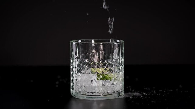 Clear liquid is poured into a glass with ice and lime over black background, slow motion cinematic clip.