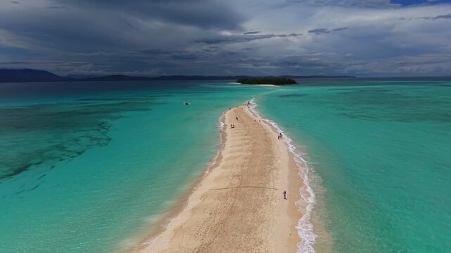 Aerial view shows a long sandy sandbar stretching between turquoise waters. People walk along the beach, enjoying the scenic view. Dark clouds loom overhead, adding drama to the scene