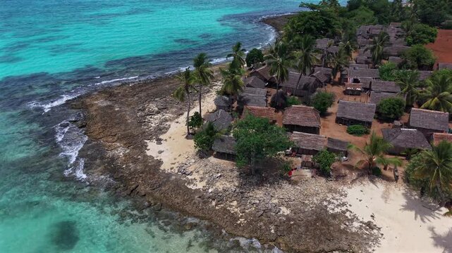 Aerial view of a tropical island with turquoise waters. Coconut palms line sandy beaches and wooden huts. People relax on shore under bright sun. Scene ideal for travel, vacation, or resort promotions