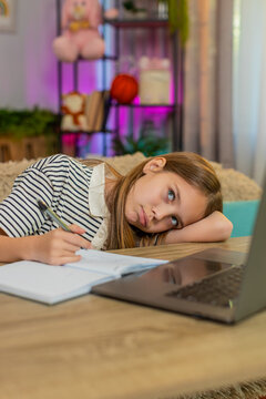 Young child girl sitting at table on home sofa, feeling tired lazy, doing homework with laptop and notebook. School kid slowly writes in exercise book struggling to stay focused during online learning