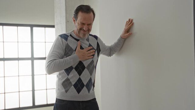 Middle aged man clutch chest for heartattack, hand braced on wall beside large window and concrete column in building; medical emergency distress.