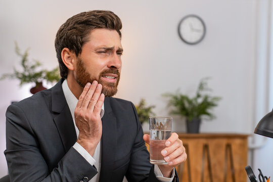 Middle-aged businessman at home office drinks cold water feels sharp tooth pain and searches dentist on smartphone. Freelancer guy at table presses cheek sighs hoping to book urgent clinic visit soon.