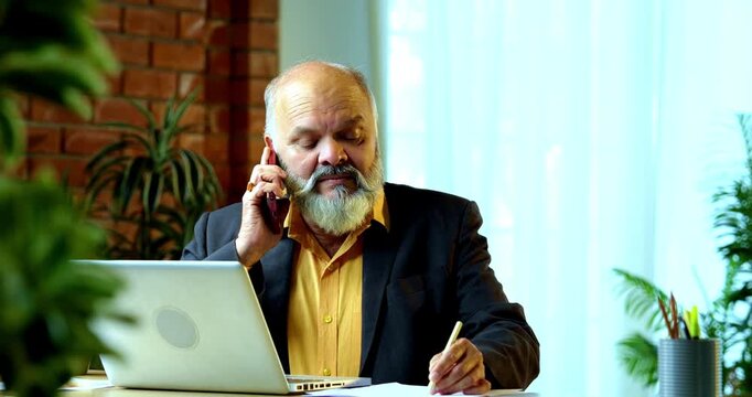 Indian senior businessman or corporate executive multitasking by taking handwritten notes while speaking on a smartphone and working on a laptop at his desk in a cozy office with moody lighting.