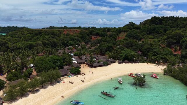Aerial view of a tropical beach with clear turquoise water. Several wooden boats float near the shore. Small thatched-roof huts line the sandy beach. People stroll along the beach