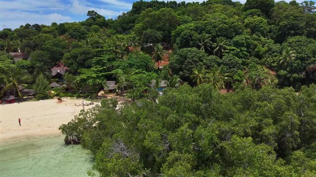 Aerial view of tropical island with white sand beach. Lush green trees cover rolling hills. Turquoise water laps shore with small boats. Few people relax near water or stroll on beach