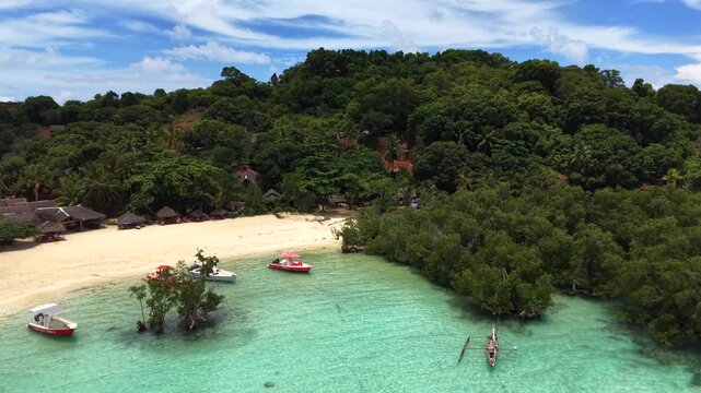 Aerial view of tropical beach with turquoise water. Small boats float near white sand shore. Lush green trees cover surrounding hills. People relax and swim in calm waters. Ideal for travel, vacation