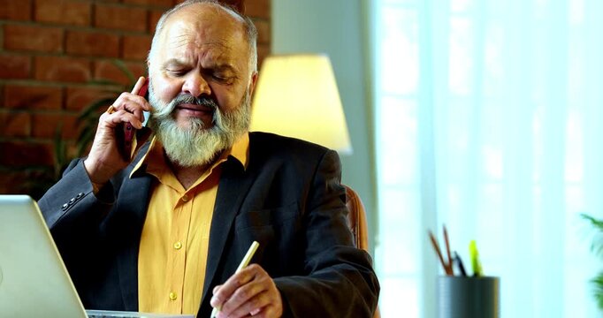 Indian senior businessman or corporate executive multitasking by taking handwritten notes while speaking on a smartphone and working on a laptop at his desk in a cozy office with moody lighting.