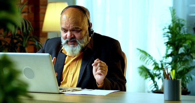 Indian senior businessman or corporate executive using a headset with a microphone for a professional discussion while working on his laptop at a desk in a cozy office with moody lighting setup.