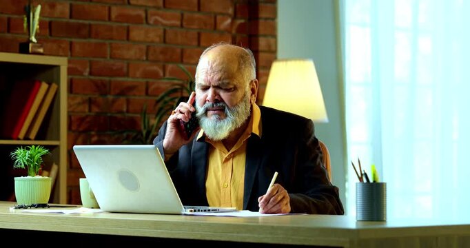 Indian senior businessman or corporate executive multitasking by taking handwritten notes while speaking on a smartphone and working on a laptop at his desk in a cozy office with moody lighting.