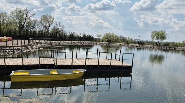 A yellow rowboat floats on a calm pond next to a wooden dock with metal railings. Lush green trees line the shore under a bright, cloudy sky, reflecting clearly in the water.