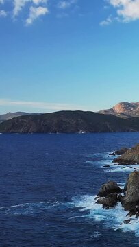 Waves crash against rugged rocks along a scenic coastline with distant hills under a clear blue sky, near Cargese, Corsica.