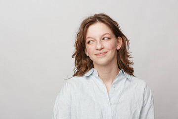 Fototapeta premium Young woman with wavy brown hair wearing a white shirt looks sideways with a slight smile on a light grey background. Concept of casual style, natural beauty, and positive mood.