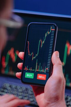 Over the shoulder view of a trader closely examining cryptocurrency charts on a smartphone via a trading app, with stock market data displayed on a laptop in the background.