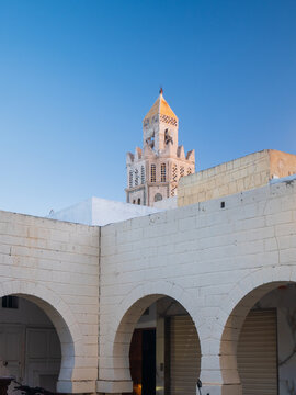 Mosque minaret rising above white arches in Sousse, Tunisia, low angle view of traditional courtyard architecture under clear blue sky, highlighting Islamic design and urban heritage.