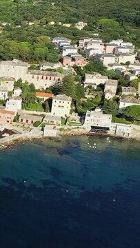 Scenic aerial shot of Erbalunga village with traditional Mediterranean houses along the clear blue coastline, surrounded by lush greenery.