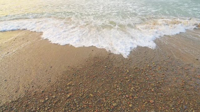 Gentle sea waves reaching sandy shore with colorful pebbles forming natural coastal texture and pattern