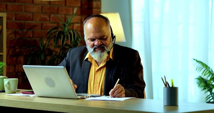 Indian senior businessman or executive wearing a headset with a microphone, taking handwritten notes while actively working on his laptop at a desk in a cozy office with professional moody lighting.