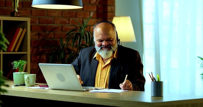 Indian senior businessman or executive wearing a headset with a microphone, taking handwritten notes while actively working on his laptop at a desk in a cozy office with professional moody lighting.