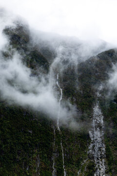 View of a misty mountain face, shrouded in low-hanging clouds, with a thin waterfall cascading down the dark, forested slopes, Milford Sound, Southland Region, New Zealand.
