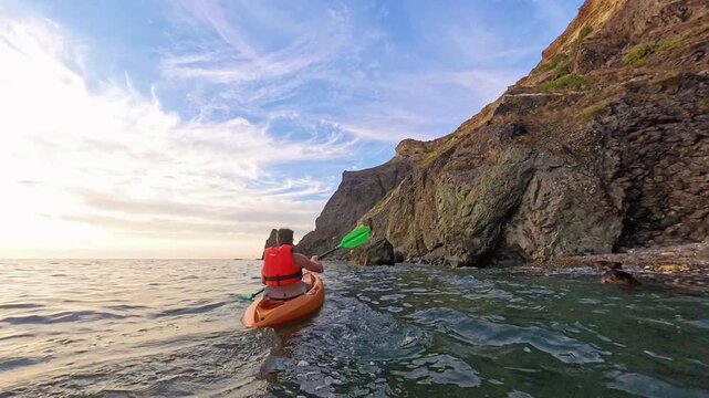 Kayak ocean paddling man in a red life vest maneuvers an orange boat along rocky cliffs and reaches for a floating balloon