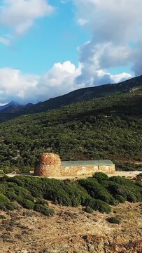 A rustic stone building with a round tower stands amidst lush green hills in the Delta du Fango, Corsica, under a partly cloudy sky.